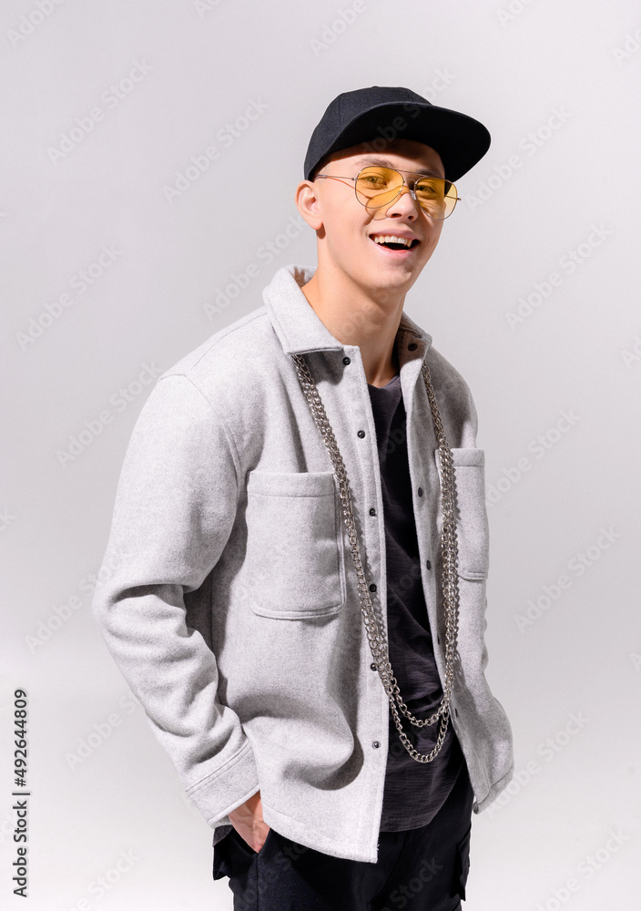 Portrait of male hipster, which wearing in casual apparel, sunglasses and cap on head, keeping hands in pockets of jeans, smiling and looking at camera while standing on white background in studio Portrait of male hipster, which wearing in casual apparel, sunglasses and cap on head, keeping hands in pockets of jeans, smiling and looking at camera while standing on white background in studio