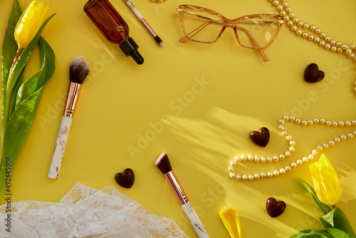 Pastel feminine flat lay with pearls, makeup brushes, glasses, oil, serum dropper and chocolate hearts. Yellow tulips with trendy shadows. Beauty card concept. Copy space photo