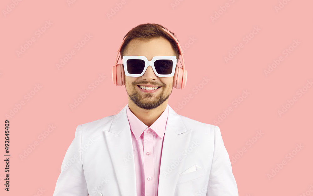 Portrait of smiling young man in modern stereo headphones isolated on pink background. Close up of cheerful guy in white jacket and sunglasses listening to music with modern wireless headphones.