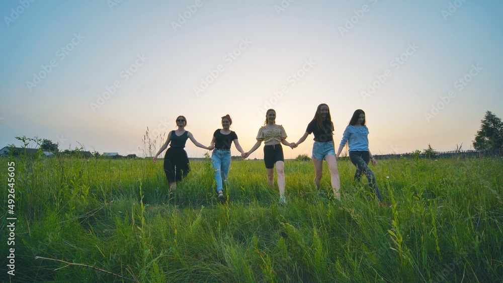 Girls friends go hand in hand at sunset across the field.