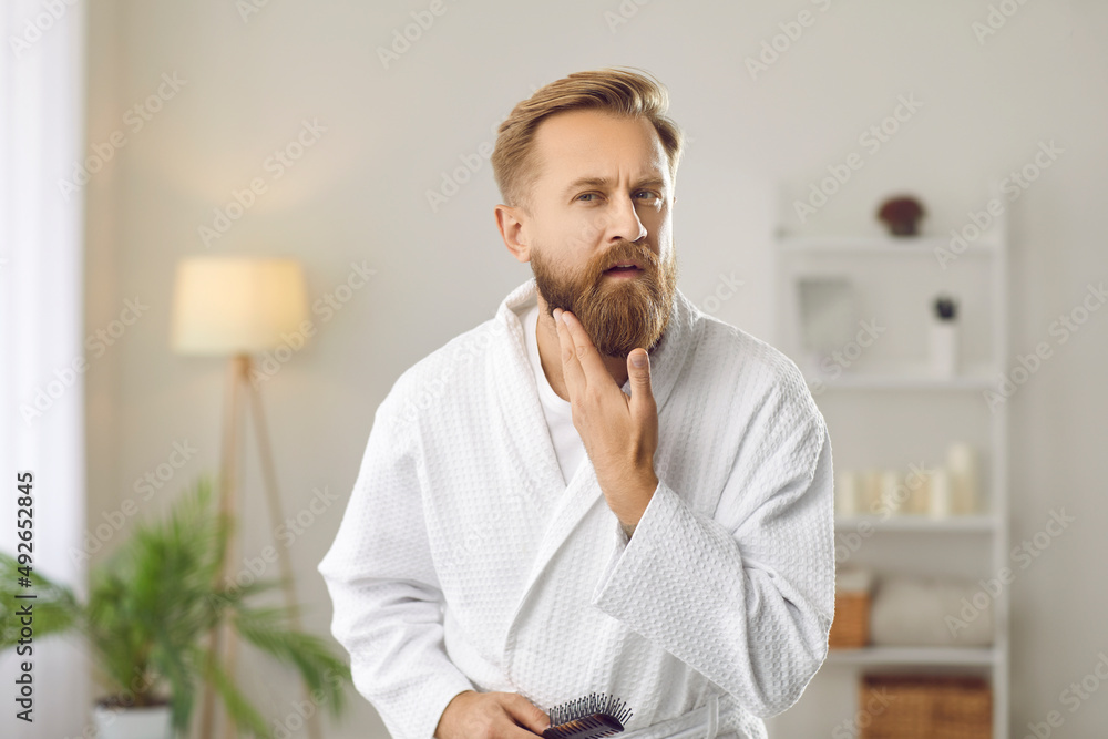 Fixing beard style. Morning preparation of attractive adult man in white bathrobe who fixes his beard looking into camera. Ad of male haircare cosmetics products. Concept of men's care. Fixing beard style. Morning preparation of attractive adult man in white bathrobe who fixes his beard looking into camera. Ad of male haircare cosmetics products. Concept of men's care.