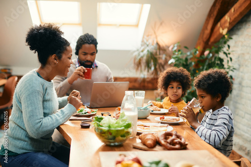 Happy black mother talks to her children during breakfast at dining table while father is working on laptop.