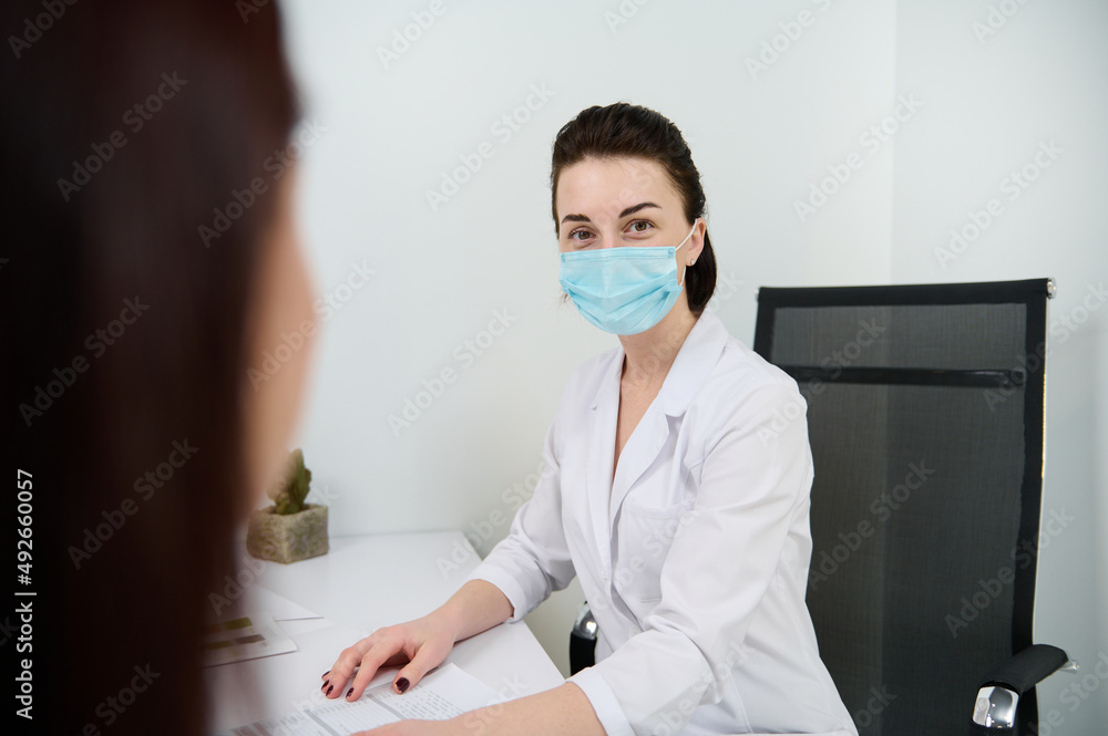Confident portrait of a brunette Caucasian woman, female doctor in medical mask looking at camera while consulting a patient in the modern minimalist medical office in a clinic with white interior