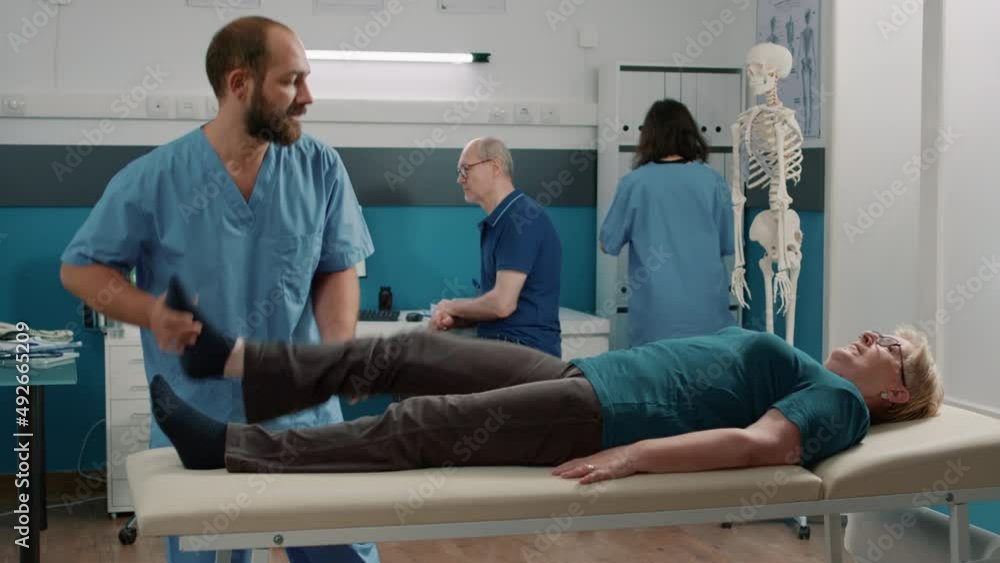 Elder person raising legs to stretch muscles in osteopathy cabinet, receiving support from male physiotherapist. Medic helping with stretching procedure exercise at rehabilitative physiotherapy. Elder person raising legs to stretch muscles in osteopathy cabinet, receiving support from male physiotherapist. Medic helping with stretching procedure exercise at rehabilitative physiotherapy.