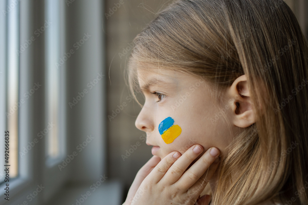 the child looking at the window with the flag of Ukraine. State support in war. Hope for the peace. Children's tears. the child looking at the window with the flag of Ukraine. State support in war. Hope for the peace. Children's tears.