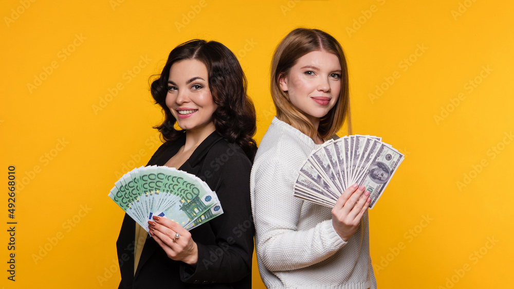 Two Delighted Girls Rejoicing Win While Holding Fans of Cash Money Dollars and Euro. Young Women Showing Bunch Money Banknotes and Looking at Camera on Yellow Background. Rich Girlfriends. Two Delighted Girls Rejoicing Win While Holding Fans of Cash Money Dollars and Euro. Young Women Showing Bunch Money Banknotes and Looking at Camera on Yellow Background. Rich Girlfriends.