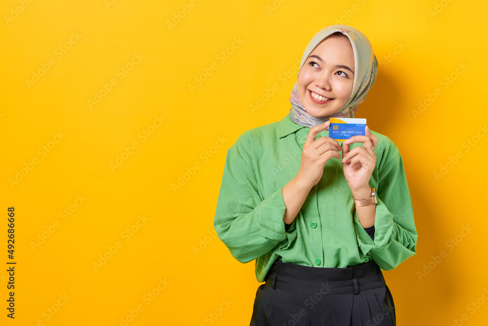 Thoughtful young Asian woman in a green shirt holding credit card and looking away on yellow background Thoughtful young Asian woman in a green shirt holding credit card and looking away on yellow background