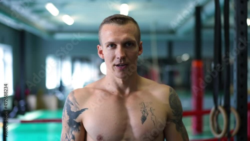 Confident tattooed young sportsman looking at camera breathing heavily standing in gym indoors. Front view portrait of muscular Caucasian man posing in slow motion indoors. Live camera zoom out