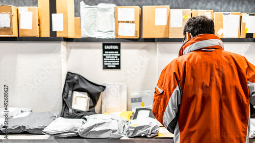 Man standing at mailroom inside apartment looking for packaging. Messenger deliver parcel or receiver concept. Guy in red uniform jacket with pile of delivering bags and boxes at distributor storage.