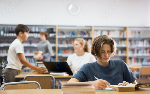 Intelligent male teenager engaged in research working with books in library