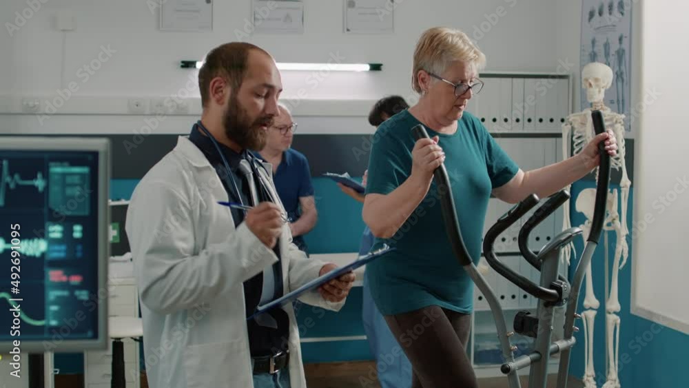 Osteopath taking notes and watching patient use stationary bicycle, doing physical exercise for muscle recovery. Treat mechanical disorders with electrical bike and heart rate monitor. Osteopath taking notes and watching patient use stationary bicycle, doing physical exercise for muscle recovery. Treat mechanical disorders with electrical bike and heart rate monitor.