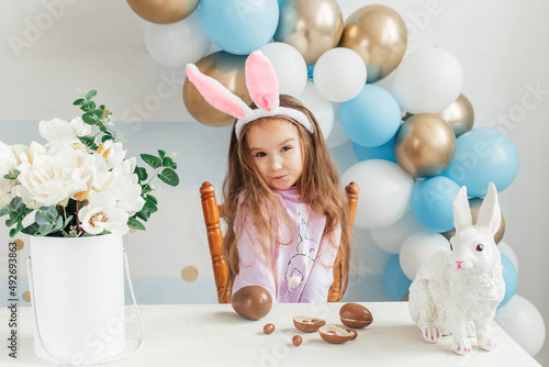 Photography Little cute girl in easter bunny ears sits at the table and plays with easter chocolate eggs