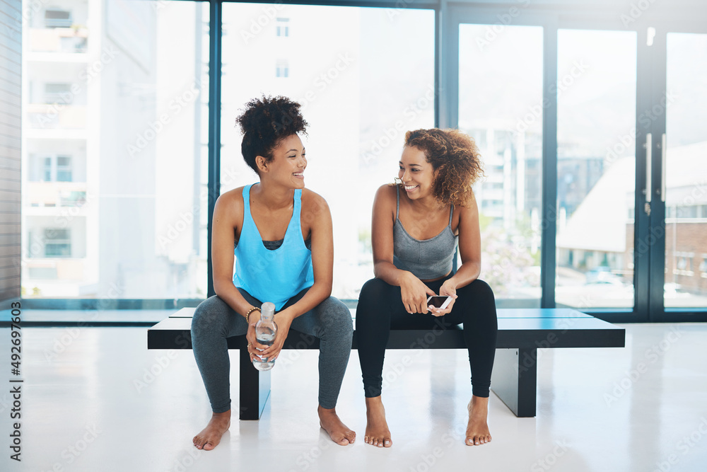 Lets take a bit of a breather. Portrait shot of two young fit women sitting down on a bench and having a chat before a yoga session inside a studio. Lets take a bit of a breather. Portrait shot of two young fit women sitting down on a bench and having a chat before a yoga session inside a studio.
