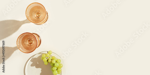 White wine glasses peach color glass and grapes fruit on beige background with dark shadow, glare from sun. Minimal summer rest concept. Dry wine in colored glassware goblets style. Creative photo