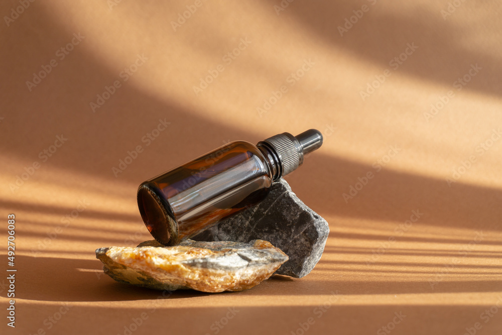 Concept of beauty, rejuvenation. Serum in an amber glass bottle with dropper lid on brown background with shadow in form of stripes. Essential oil for women's skin care on 3D granite stone stand