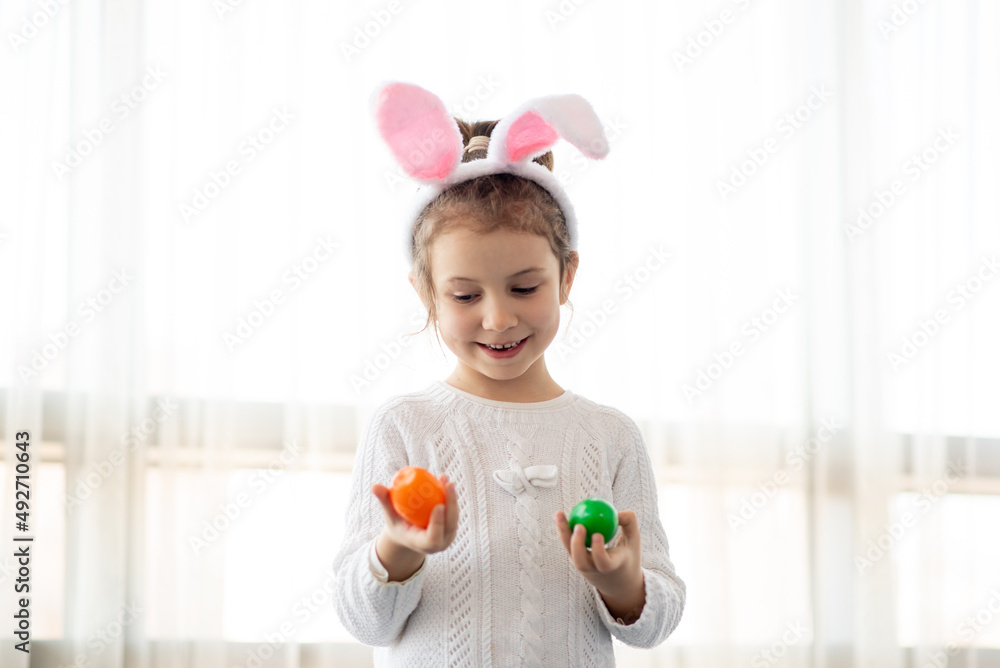A little cute girl in rabbit ears holds colorful eggs in her hands. She is wearing a white T-shirt. Easter. Preparation for the holiday. A little cute girl in rabbit ears holds colorful eggs in her hands. She is wearing a white T-shirt. Easter. Preparation for the holiday.
