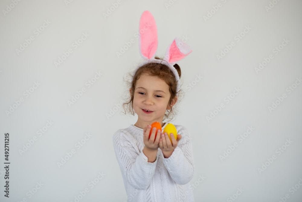 A little cute girl in rabbit ears holds colorful eggs in her hands. She is wearing a white T-shirt. Easter. Preparation for the holiday. A little cute girl in rabbit ears holds colorful eggs in her hands. She is wearing a white T-shirt. Easter. Preparation for the holiday.
