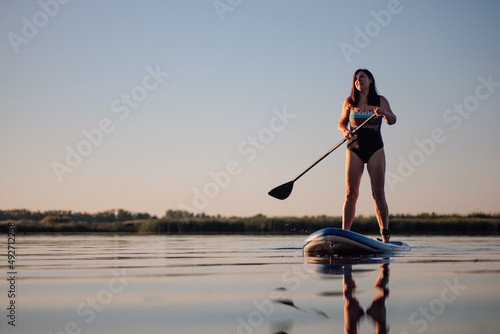 Low-angle of Caucasian woman standing on sup board with one oar in hands looking up at sky on crystal blue lake wearing summer swimming dress. Active lifestyle. 