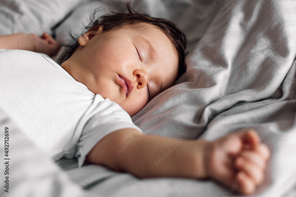 Portrait of baby in sleepwear lying under blanket on bed at home. Sleeping infant child in bedroom, see sweet dream. Grey background, soft focus, free copy space. Childcare and healthy sleep concept. Portrait of baby in sleepwear lying under blanket on bed at home. Sleeping infant child in bedroom, see sweet dream. Grey background, soft focus, free copy space. Childcare and healthy sleep concept.