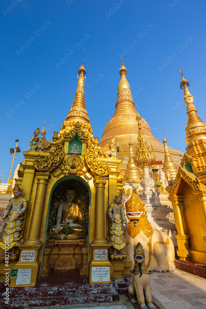 Naklejka premium Small pagodas and statues in front of the gilded Shwedagon Pagoda in Yangon, Myanmar on a sunny day.