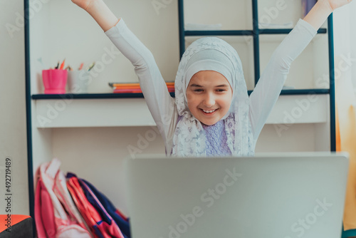 A happy Muslim student girl learning online education on laptop in the home. A smiling girl in hijab having online communication with friends on internet. Happy Muslim kid in hijab using a laptop. photo