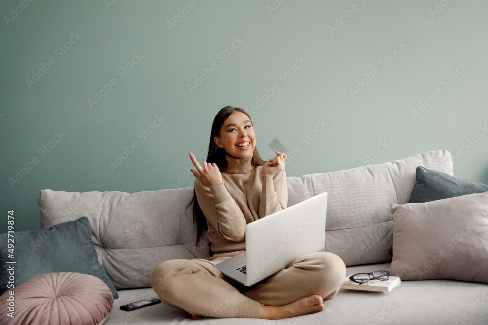 Smiling beautiful woman holding credit card and using laptop computer sitting on couch at home,. Online shopping concept. Smiling beautiful woman holding credit card and using laptop computer sitting on couch at home,. Online shopping concept.