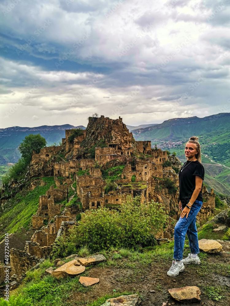 Fototapeta premium A girl on the background of Gamsutl village in the Caucasus mountains, on top of a cliff. Dagestan Russia June 2021