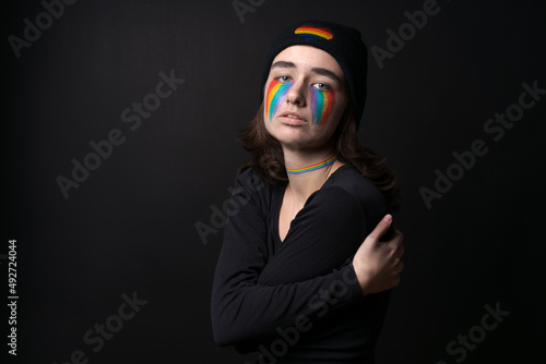 Portrait young crying woman with painted LGBT flag on her cheek. Crying with rainbow in black hat with rainbow in form heart on black background. Concept violation rights and equality of LGBT people.