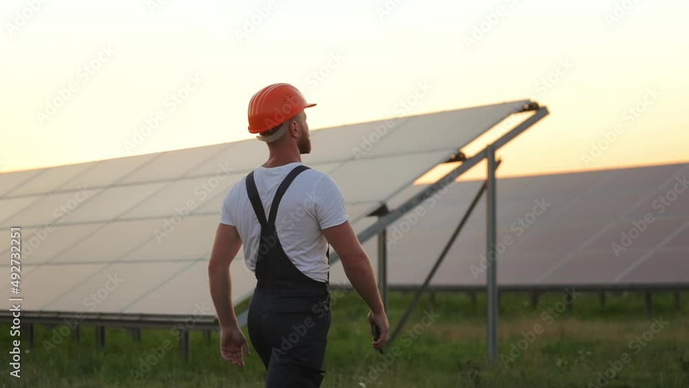 Back view of professional technician in overalls and helmet walking on solar station with modern smartphone in hands. Summer sunset on background. Production of green energy. Back view of professional technician in overalls and helmet walking on solar station with modern smartphone in hands. Summer sunset on background. Production of green energy.