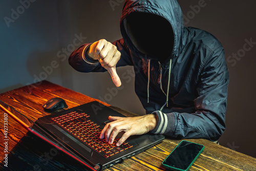 Fototapeta Naklejka Na Ścianę i Meble -  A hacker anonymous in a hood is typing on a laptop keyboard in a dark room. Cybercrime fraud and identity theft