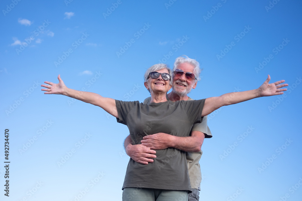 Happy senior man hugging his wife while she stands with open arms. Beautiful smiling white-haired elderly couple enjoying retirement and freedom on outdoor excursion Happy senior man hugging his wife while she stands with open arms. Beautiful smiling white-haired elderly couple enjoying retirement and freedom on outdoor excursion