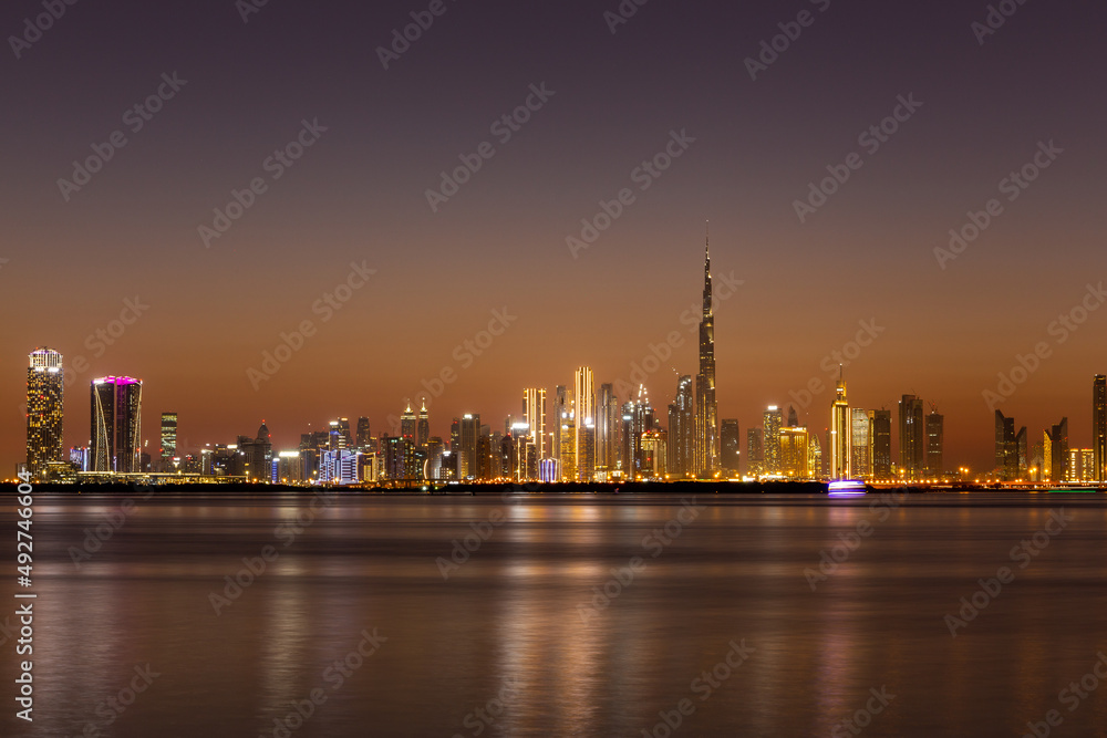 Dubai Business Bay skyline at night with colorful illuminated buildings and calm Dubai Creek water.