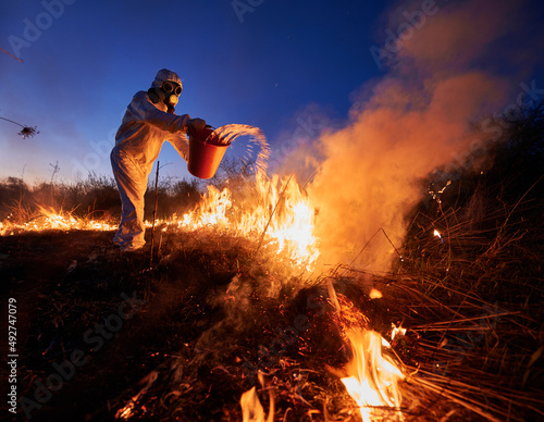 Research scientist fighting fire in field with blue night sky on background. Man in protective radiation suit and gas mask holding bucket and pouring water on burning grass with smoke.