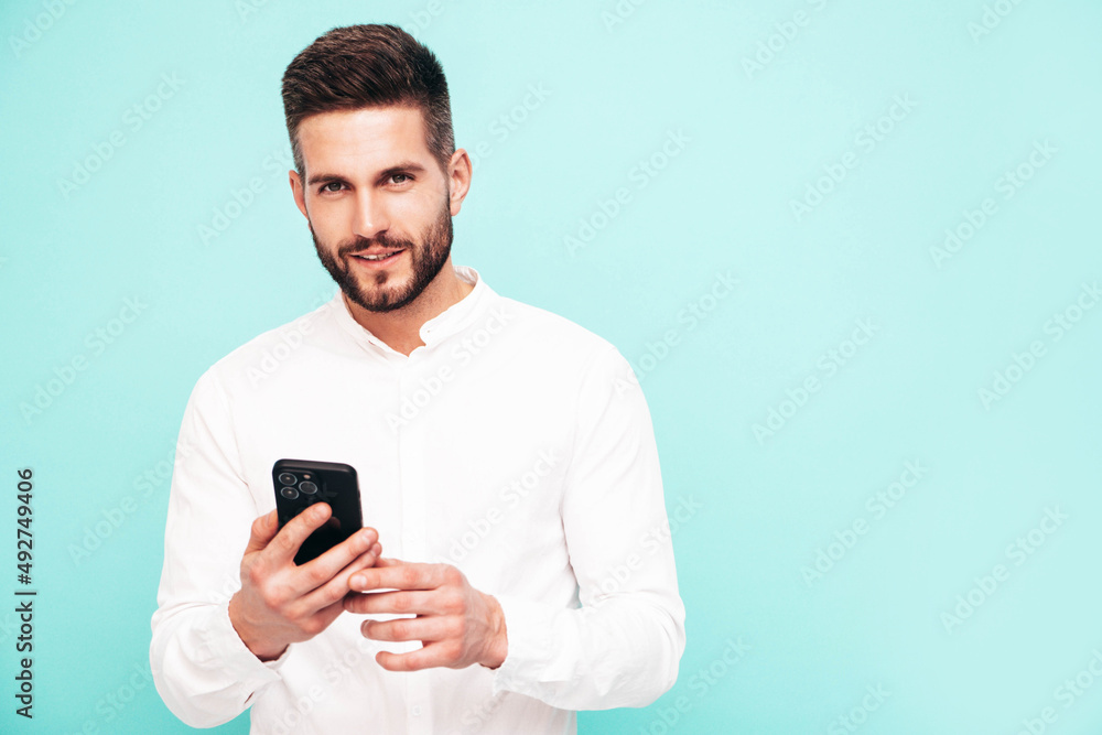 Handsome smiling model.Sexy stylish man dressed in shirt and jeans. Fashion hipster male posing near blue wall in studio. Holding smartphone. Looking at cellphone screen. Using apps Handsome smiling model.Sexy stylish man dressed in shirt and jeans. Fashion hipster male posing near blue wall in studio. Holding smartphone. Looking at cellphone screen. Using apps