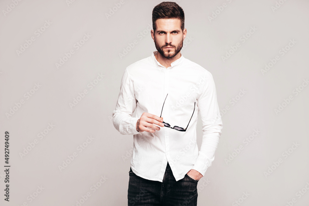 Portrait of handsome confident model. Sexy stylish man dressed in white shirt and jeans. Fashion hipster male posing in studio on grey background. Holding sunglasses. Isolated Portrait of handsome confident model. Sexy stylish man dressed in white shirt and jeans. Fashion hipster male posing in studio on grey background. Holding sunglasses. Isolated