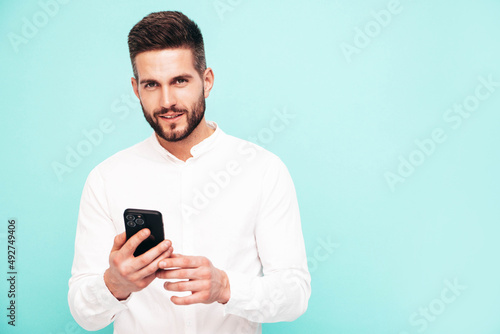 Handsome smiling model.Sexy stylish man dressed in shirt and jeans. Fashion hipster male posing near blue wall in studio. Holding smartphone. Looking at cellphone screen. Using apps