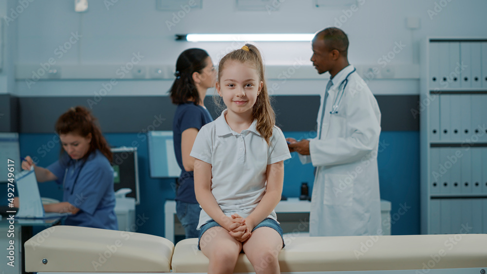 Obraz premium Portrait of little girl on bed waiting to begin checkup exam in medical cabinet with doctor. Small kid with illness looking at camera and getting ready for checkup visit appointment in office.
