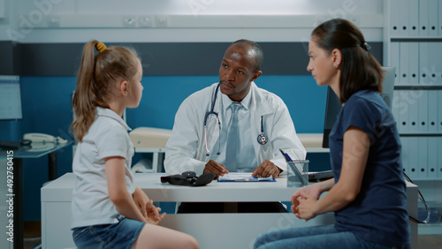 Doctor talking to patients and taking notes on computer to prepare checkup document with prescription medicine. Medic having conversation with little girl and adult in medical cabinet.