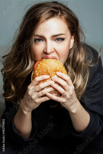 Hungry woman eats a hamburger. Beautiful blonde in gray pajamas. Junk food and unhealthy. Gray background. Vertical. Close-up.