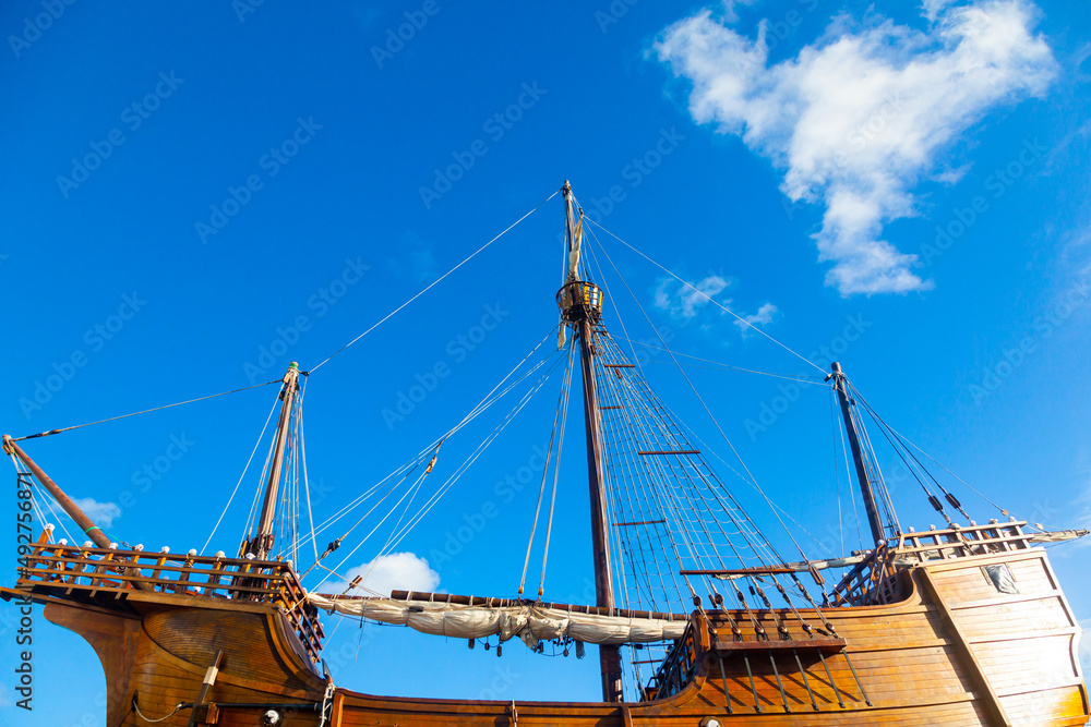 An old wooden caravel ship with folded sails against a blue sky with white clouds.