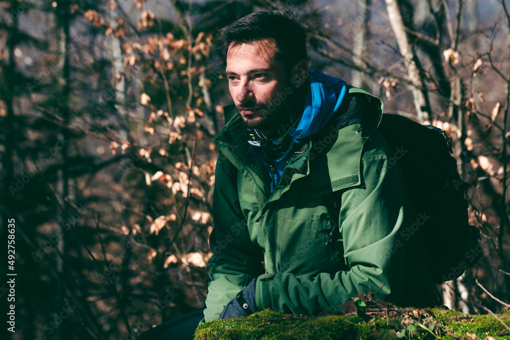 Attractive Caucasian male hiker posing in forest  Waist up shot of young bearded man with serious look on his face looking away in woodland.