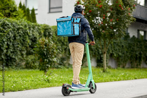 food shipping, transportation and people concept - delivery man in bike helmet with thermal insulated bag riding electric scooter on city street