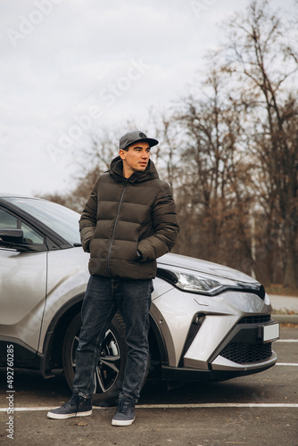 A young cheerful guy in a jacket and a cap stands near a new car photo