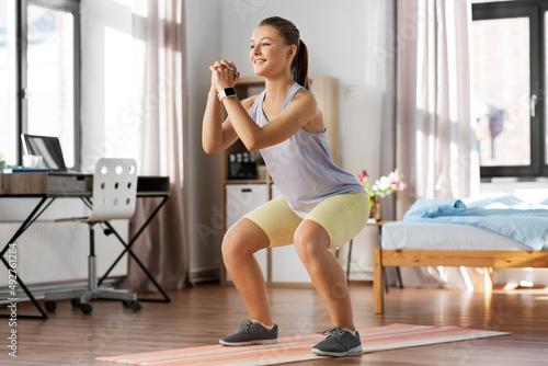 sport, fitness and healthy lifestyle concept - smiling teenage girl with smart watch exercising on yoga mat and doing squats at home