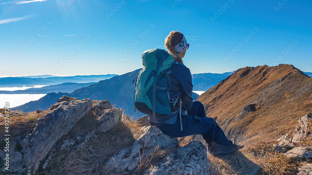 A female hiker having a break on the summit of Eisenerzer Reichenstein in Styria, Austria, Europe. The Ennstal valley is covered in clouds and fog. Hiking trail, Wanderlust, Season. Sunny day. Freedom