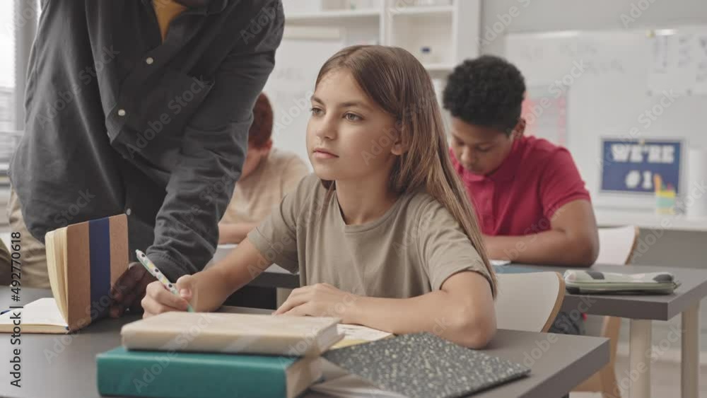 Waist up slowmo of unrecognizable African American teacher standing by desk of female pupil writing test and helping her Waist up slowmo of unrecognizable African American teacher standing by desk of female pupil writing test and helping her