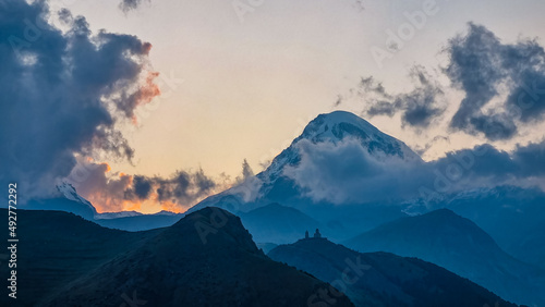 Distant view on Gergeti Trinity Church in Stepansminda, Georgia. The church is located the Greater Caucasian Mountain Range.Clouds are covering the snow-capped Mount Kazbegi in the back.Sunrise,sunset