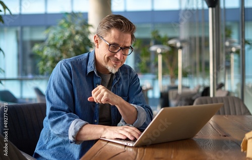 Happy middle age businessman using laptop computer in office. Older casual man working online on terrace of cafe, smiling.