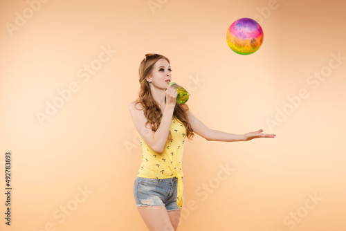 A woman in bright clothes, a yellow T-shirt and denim shorts, sunglasses holds a ball and cold lemonade in her hands, on an isolated background © Вячеслав Косько