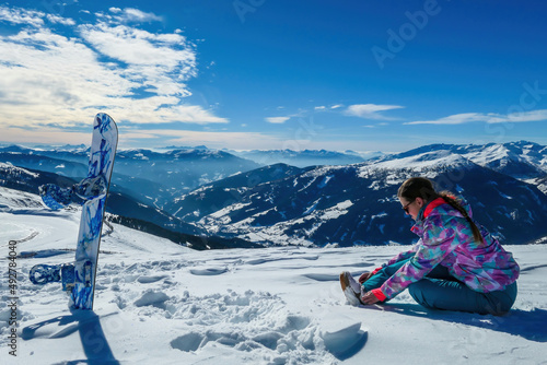A woman sitting on the snow on top of Katschberg in Austria. She is happy and joyful. Panoramic view on the surrounding mountains. Winter wonderland. Bright and sunny winter day. Love and happiness